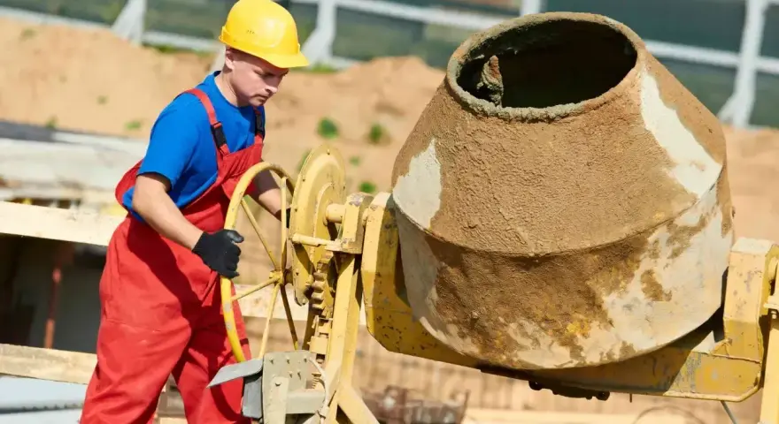 Robotnik w żółtym kasku i czerwonym ogrodniczkach obsługuje betoniarkę. Mieszanka cementowa wygląda na gładką, bez widocznego żwiru, co sugeruje specyficzne proporcje betonu.