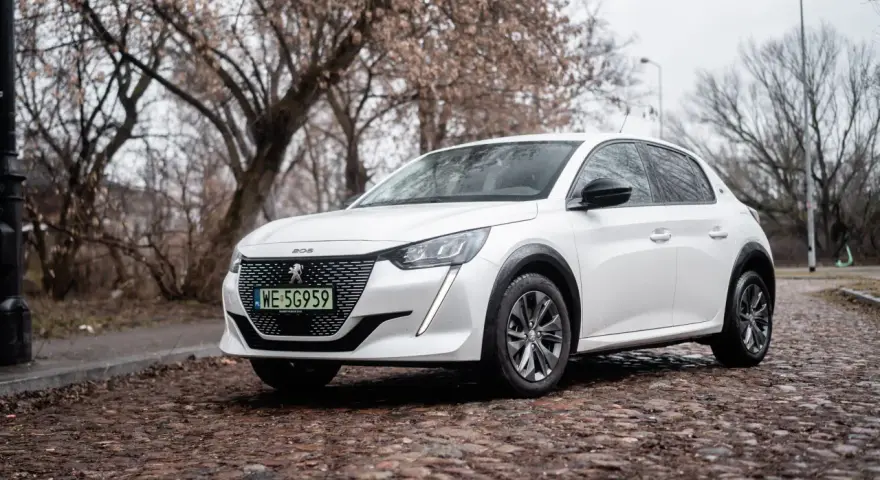 Biały Peugeot e-208 na brukowanej ulicy, z drzewami w tle.