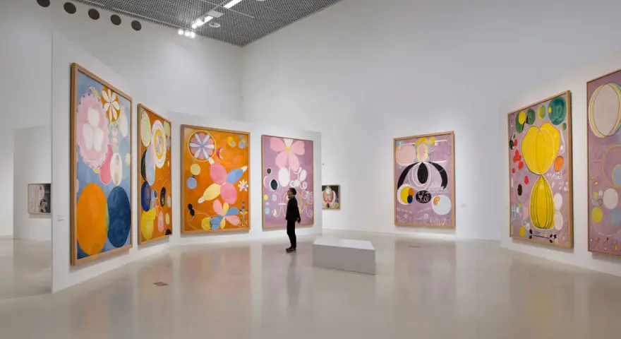 En person betraktar en samling färgstarka, abstrakta målningar av Hilma af Klint i ett ljust galleri.