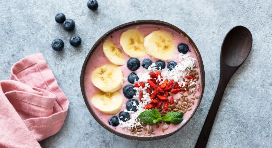 Kolorowy smoothie bowl z bananem, jagodami i kokosem, przypominający wyglądem tradycyjny kisiel. Obok leży drewniana łyżka i różowa serwetka.