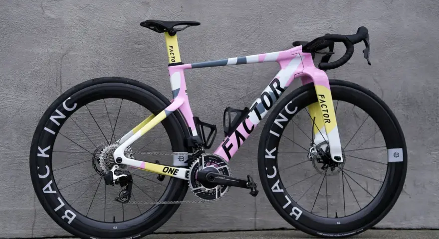 A pink, yellow, and gray Factor road bike with black wheels against a gray wall.