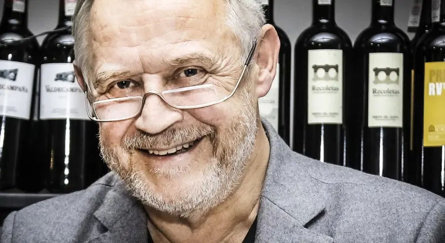 Marek Kondrat, z uśmiechem i okularami, na tle butelek wina. Jego majątek to nie tylko sukcesy zawodowe, ale i pasja do trunków.