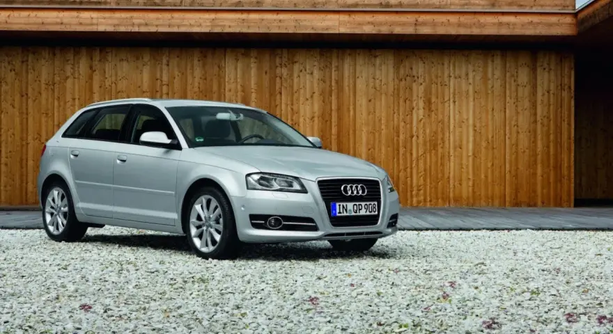 Silberner Audi A3 Baujahr 2008 steht auf einem Kiesparkplatz vor einer Holzwand.
