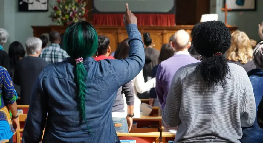 A woman with green hair raises her hand, perhaps inspired by the lyrics on screen, offering chant ideas for the congregation.