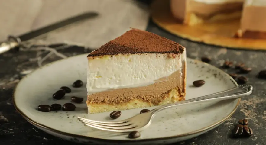 Pyszny sernik tiramisu z warstwami kremu i biszkoptu, posypany kakao. Na talerzu ziarna kawy i widelec.