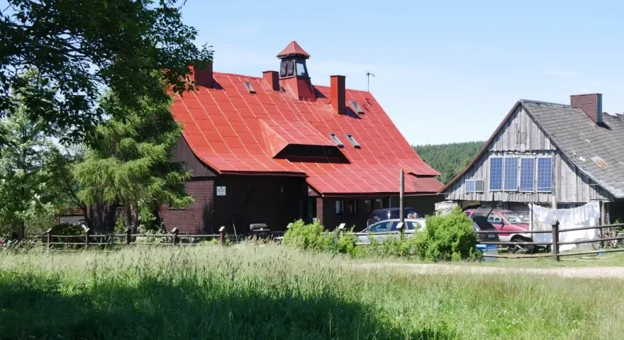 Górzyste krajobrazy, gdzie króluje chatka górzystów z czerwoną dachem. Wokół zieleń i kilka samochodów.