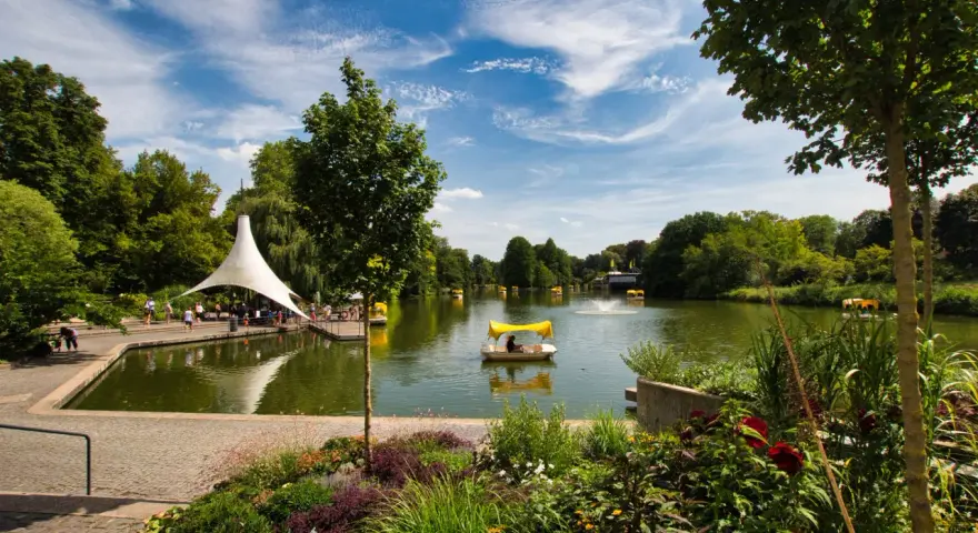 Ein sonniger Tag am Badesee Mannheim. Mehrere Tretboote mit gelben Verdecken gleiten über das Wasser, umgeben von üppigem Grün und blühenden Blumen.