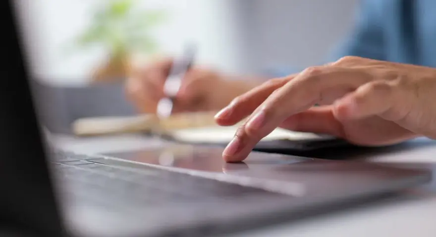 Dłoń na touchpadzie laptopa, w tle notatnik i długopis.