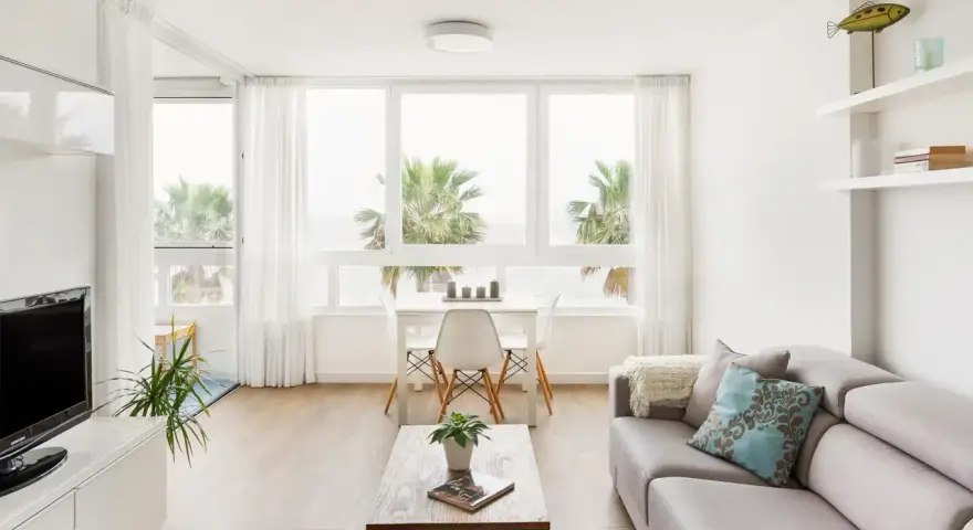 Un luminoso salón estilo nórdico con sofá gris, mesa de centro de madera y grandes ventanales con vistas a palmeras.