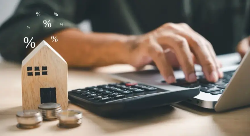 Drewniany domek, monety i kalkulator symbolizują planowanie finansowe. To idealny obraz dla osób szukających mieszkania na start.