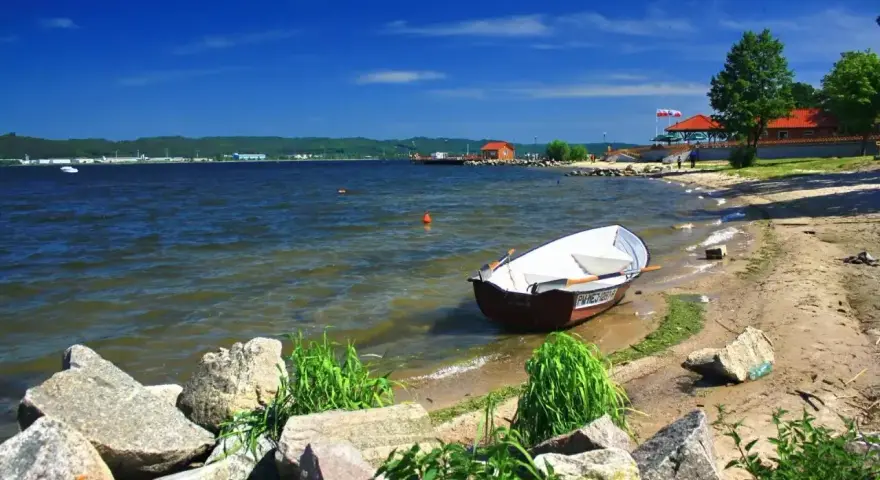 Łódka zacumowana przy brzegu jeziora żarnowieckiego. W tle budynki i zielone wzgórza.