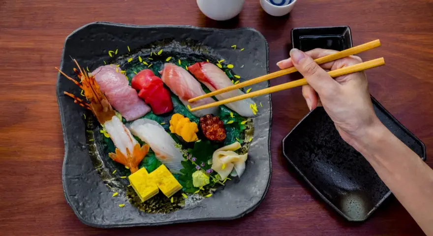 Wykwintne jedzenie w Japonii: ręka z pałeczkami sięga po świeże sushi i sashimi, obok sos i sake.