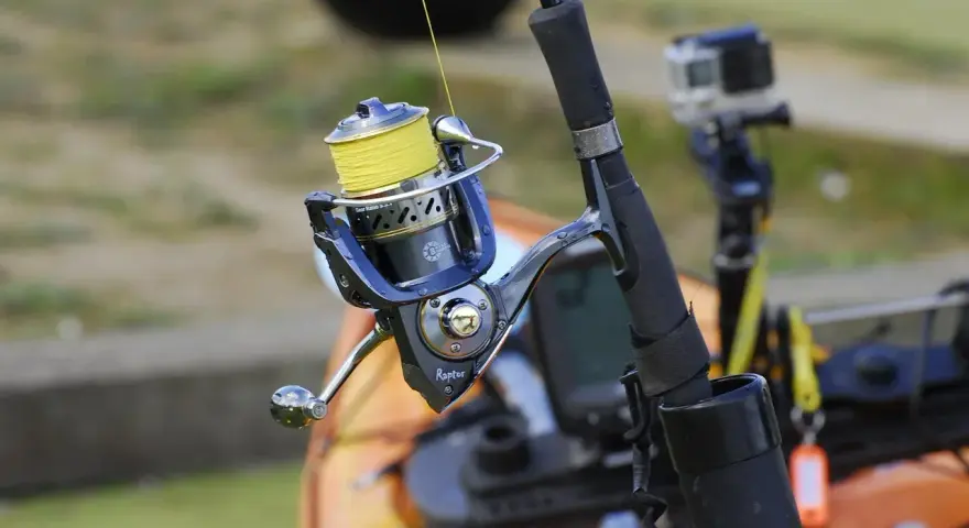 Niebieski kołowrotek spinningowy z żółtą żyłką, gotowy do akcji. W tle widać kajak i kamerę GoPro.