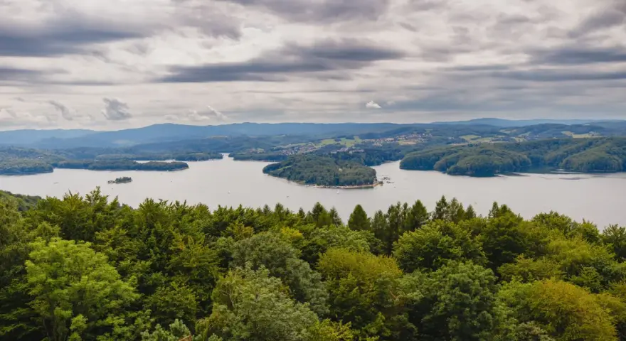 Widok na jezioro otoczone lasami, z górami w tle. Na pierwszym planie bujna zieleń drzew, a dalej spokojna tafla wody i zabudowania. Nad wszystkim chmury, jakby biała góra na niebie.