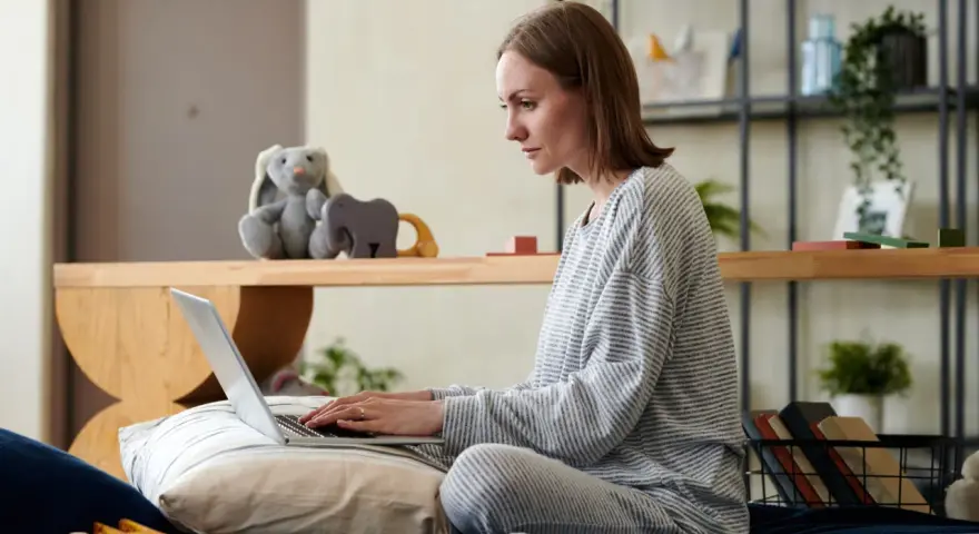 Kobieta w piżamie pisze na laptopie, przygotowując wniosek o urlop macierzyński. Obok pluszowy królik i zabawki.