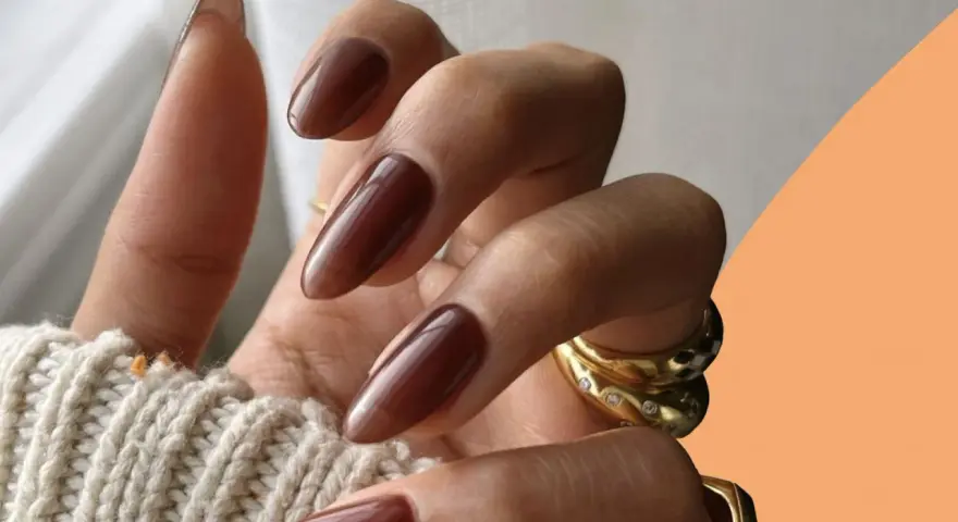 Mani con unghie autunnali chiare color cioccolato, lunghe e affusolate, adornate da anelli dorati.