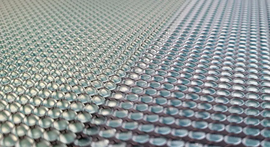 Close-up of a 3d printed lens array, with rows of tiny, clear, dome-shaped lenses reflecting light.