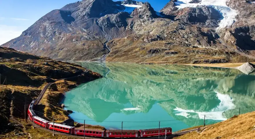 Czerwony pociąg wije się wzdłuż turkusowego jeziora u podnóża alpejskich szczytów. Idealne miejsce na trekking w alpach.