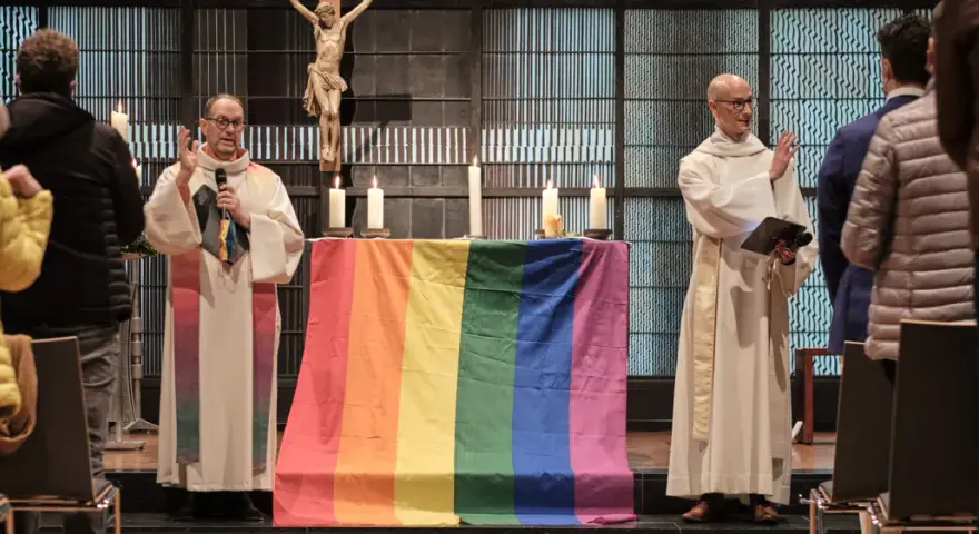 Zwei Geistliche stehen vor einem Altar mit einer Regenbogenfahne. Die Kirche feiert die gleichgeschlechtliche Ehe.