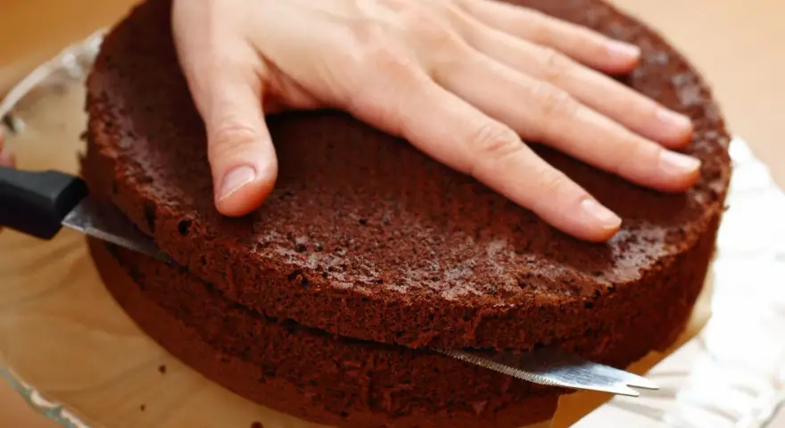 Eine Hand hält einen Schokoladenkuchen, während ein Messer ihn waagerecht durchschneidet, um den tortenboden zu teilen.