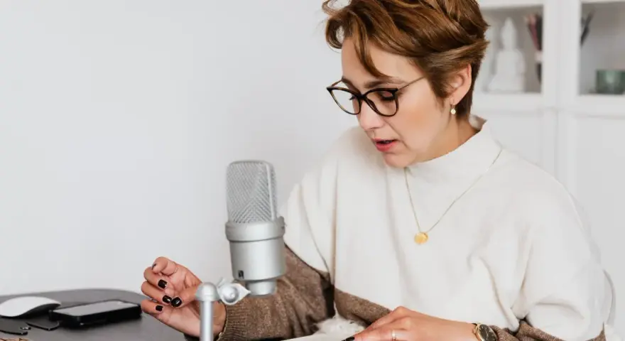 Mujer con gafas lee un libro frente a un micrófono, aprendiendo como escribir mejor.