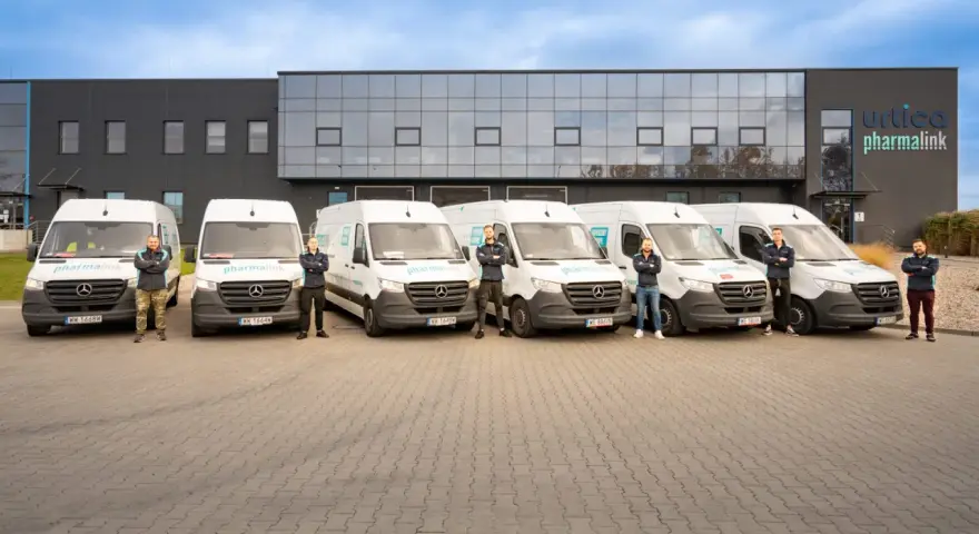 Sześciu mężczyzn w uniformach stoi przed rzędem białych vanów z logo "pharmalink". To zespół odpowiedzialny za transport leków, profesjonalny przewoźnik.