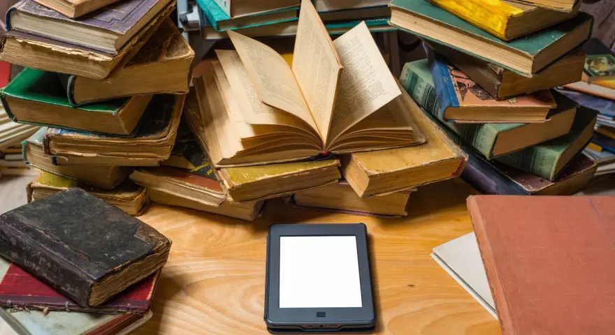 Stosy starych książek i nowoczesny ebook na drewnianym blacie. Wybór między tradycją a technologią.