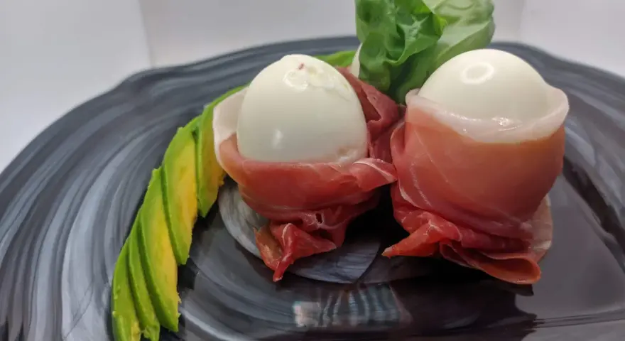 Due uova di quaglia sode avvolte in prosciutto crudo, servite con fette di avocado e foglie verdi su un piatto scuro.