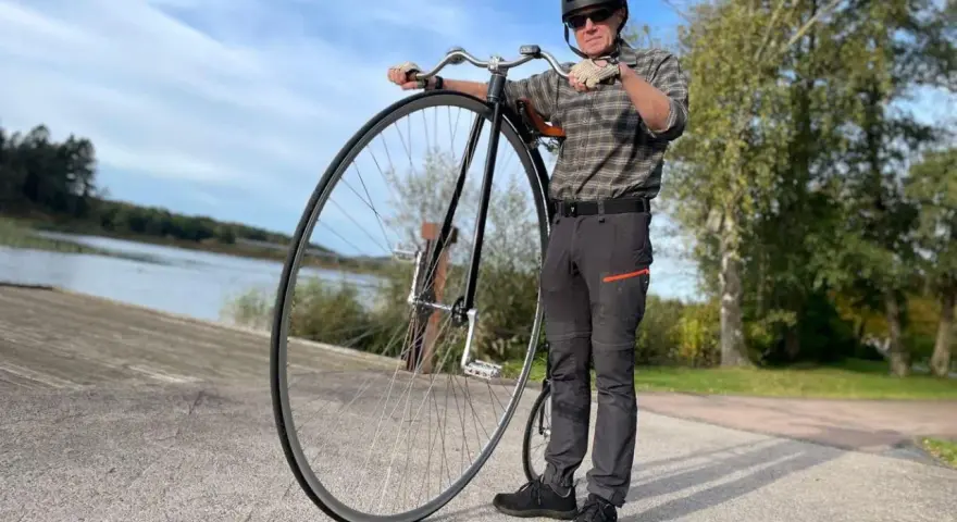 Man med hjälm och solglasögon står bredvid en cykel med stort framhjul vid en sjö.
