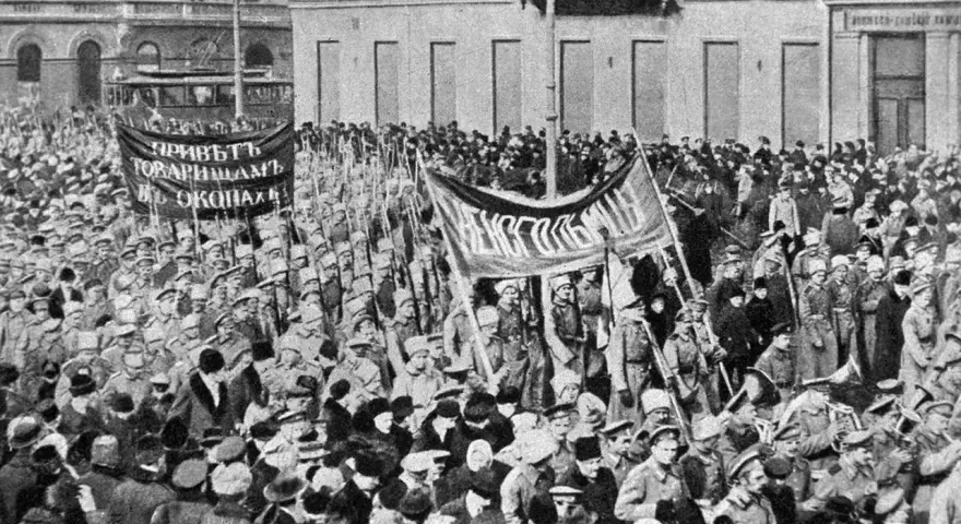 Multitud con pancartas y uniformes militares marchando en una calle, con edificios al fondo.