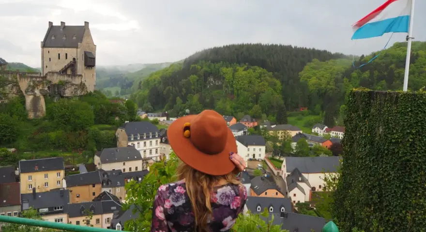 Mujer con sombrero marrón admira un castillo y un pueblo, con la bandera de Luxemburgo ondeando. ¡Un paisaje digno de un buen queso Muenster!