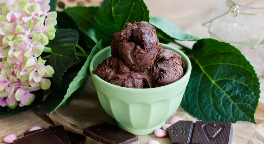 Ein grüner Napf mit zartbitter schokoladeneis, umgeben von Schokoladentafeln, rosa Bonbons und einer Hortensienblüte.