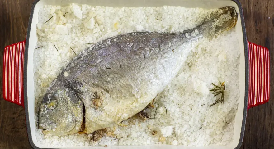 Orata al sale, intera e dorata, adagiata su un letto di sale grosso e rosmarino in una teglia rossa.