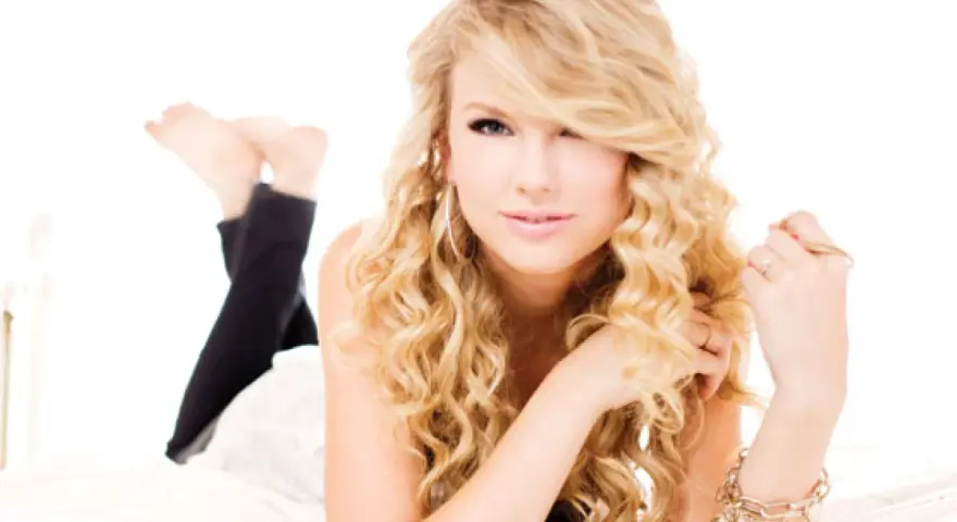 Taylor Swift w stylu "Speak Now" z burzą blond loków, w czarnych legginsach, leży na łóżku.