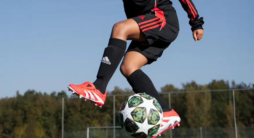 Barn i luften med fotboll. Svarta strumpor med Adidas-logga och benskydd fotboll barn storlek. Orange fotbollsskor med vita detaljer.