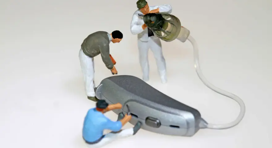 Miniaturowi pracownicy zajmują się naprawą aparatów słuchowych. Jeden z nich przykręca element, drugi czyści słuchawkę, a trzeci pracuje nad aparatem.