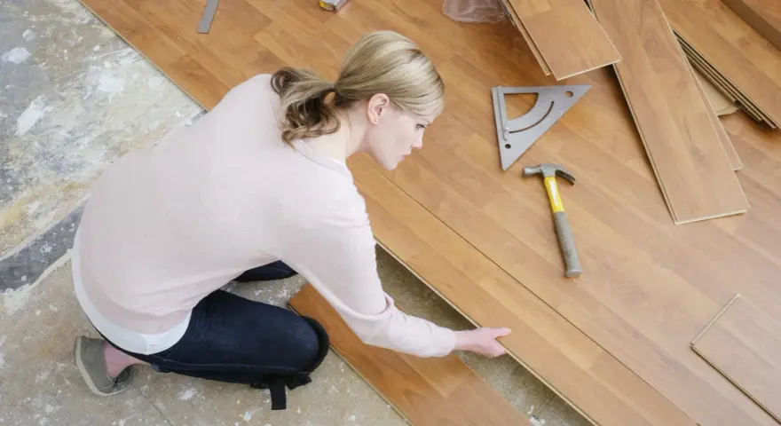 Frau kniet und verlegt Laminat. Werkzeuge wie Hammer und Winkel liegen bereit.