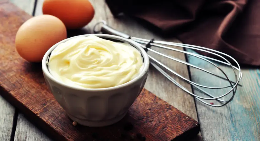 Un bol de mayonnaise onctueuse, des œufs et un fouet, prêts à faire une mayonnaise maison.