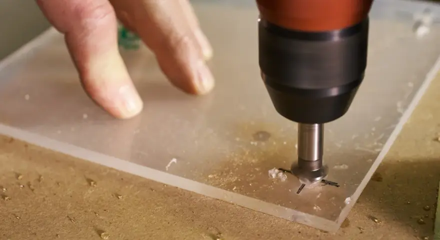 A drill bit is used to drill through acrylic. A hand guides the acrylic sheet as the drill creates a hole, showing it's possible to drill through acrylic.