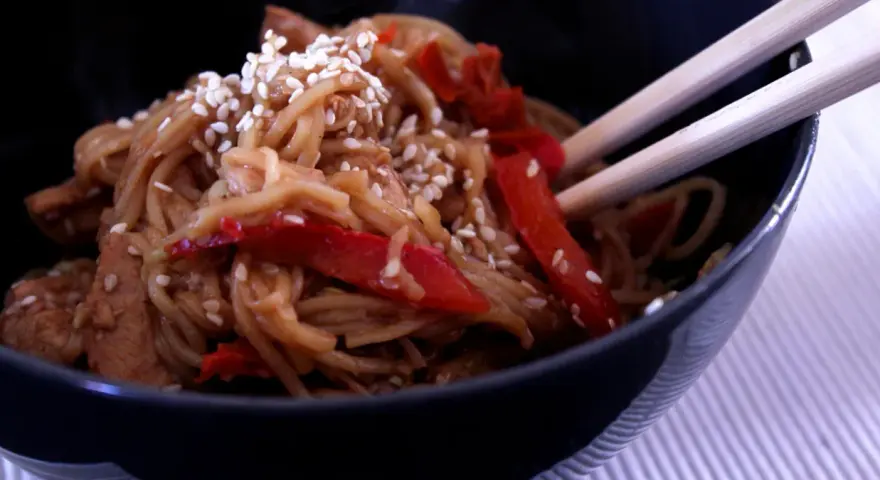Miseczka pełna aromatycznego makaronu teriyaki z kawałkami kurczaka i papryką, posypana sezamem.