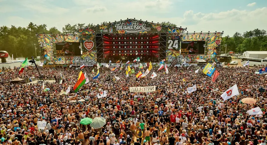 Tłum ludzi na festiwalu Poland Rock, gdzie jest scena i ekrany. Wszędzie flagi i słońce.