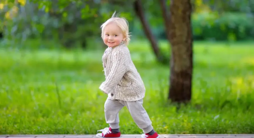 Ein fröhliches Kind, ca. 2,5 Jahre alt, mit blonden Haaren und einem Strickcardigan, läuft auf einem Weg im Park.