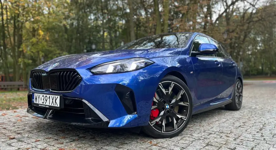 Błękitne, sportowe niemieckie auto BMW z czarnymi felgami i czerwonymi zaciskami hamulcowymi stoi na brukowanej drodze wśród jesiennych drzew.