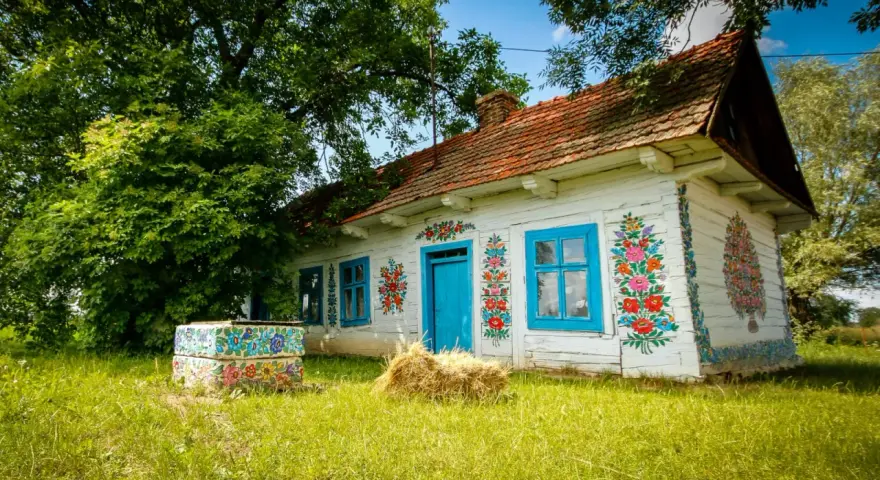 Malowany dom w Zalipiu, z niebieskimi oknami i drzwiami, ozdobiony kwiatami. Obok studnia i snop siana. Cennik wizyty w tym malowniczym miejscu jest dostępny dla zwiedzających.