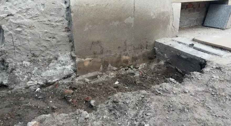 Prace przy wzmocnieniu fundamentów w starym domu. Widoczne wykopy, odsłonięta ściana i nowe elementy betonowe.