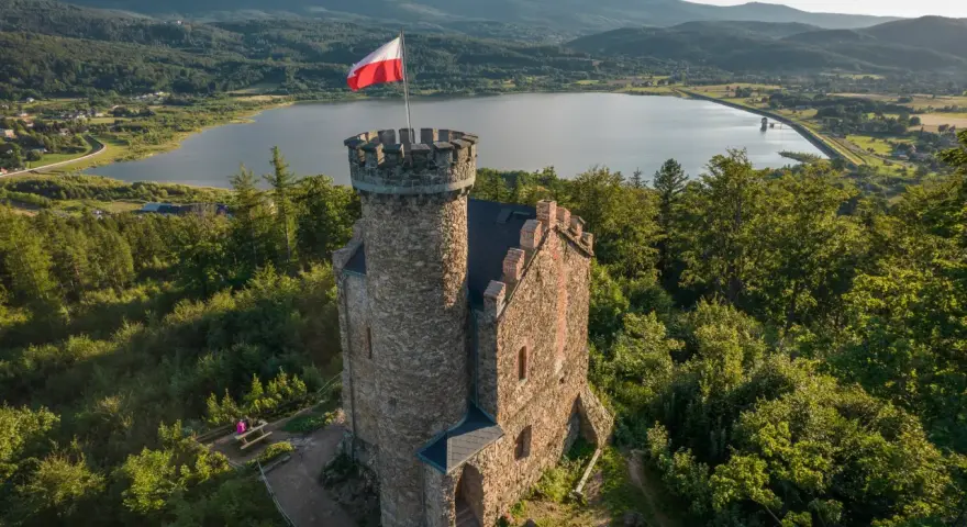 Kamienny zamek henryka z polską flagą na wieży, otoczony lasem, z jeziorem i górami w tle.
