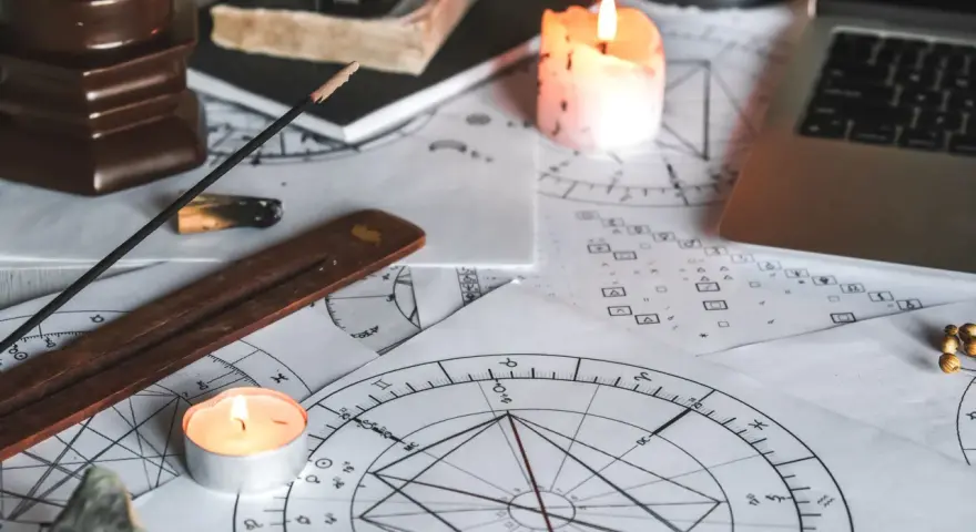 Na stole leżą wykresy astrologiczne, świece, kadzidełko i laptop. Widać symbolikę wskazującą na ascendent w bliźniętach.