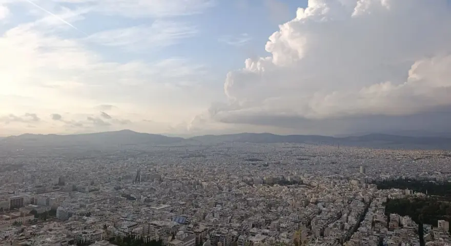 Rozległa panorama Aten z wzgórza Lycabettus. Miasto rozciąga się aż po horyzont, a nad nim piętrzą się chmury.