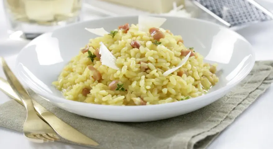 Un cremoso arroz a la milanesa, adornado con láminas de queso parmesano y hierbas frescas, servido en un plato blanco.