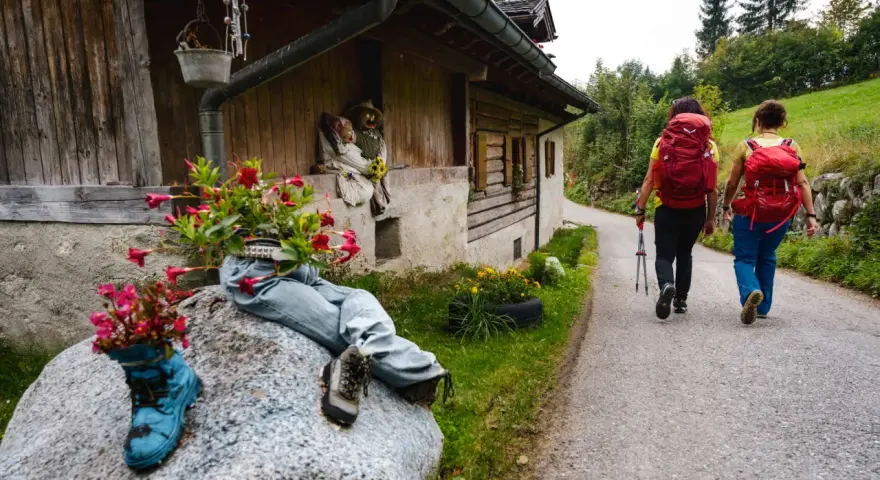 Due escursioniste affrontano il cammino San Vili, con zaini rossi e bastoncini. La strada, tra case rurali e verde, suggerisce una certa difficoltà.